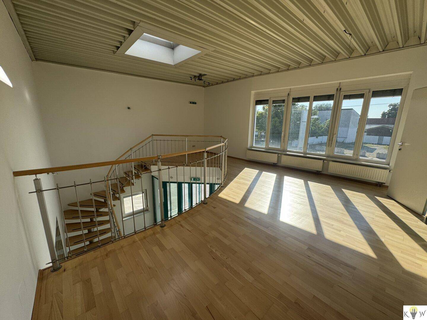Heller Büroraum im Obergeschoss mit Holzparkett, großen Fenstern und Blick auf die Treppe.