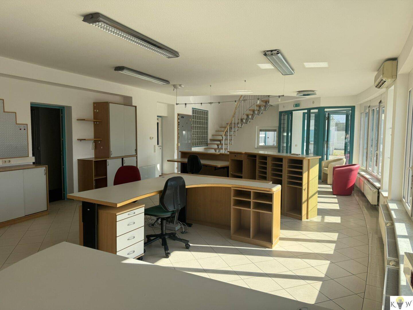 Geräumiger Bürobereich mit mehreren Arbeitsplätzen, großen Fenstern und einer Treppe zum Obergeschoss.