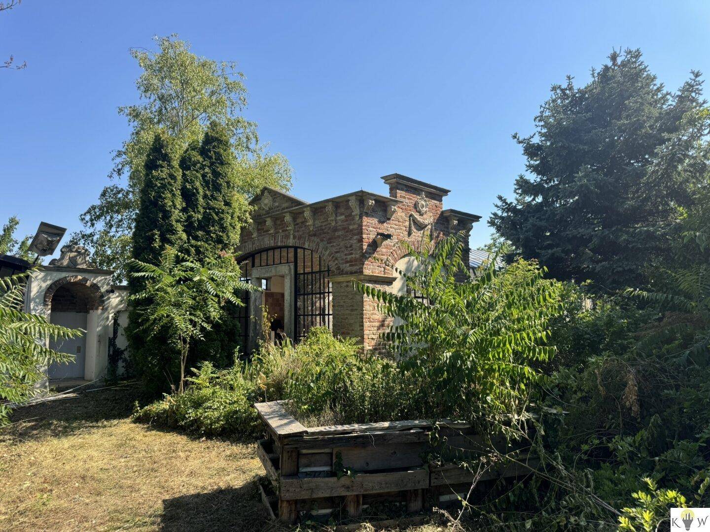 Altes Backsteingebäude mit verzierten Details, umgeben von einem verwilderten Gartenbereich.