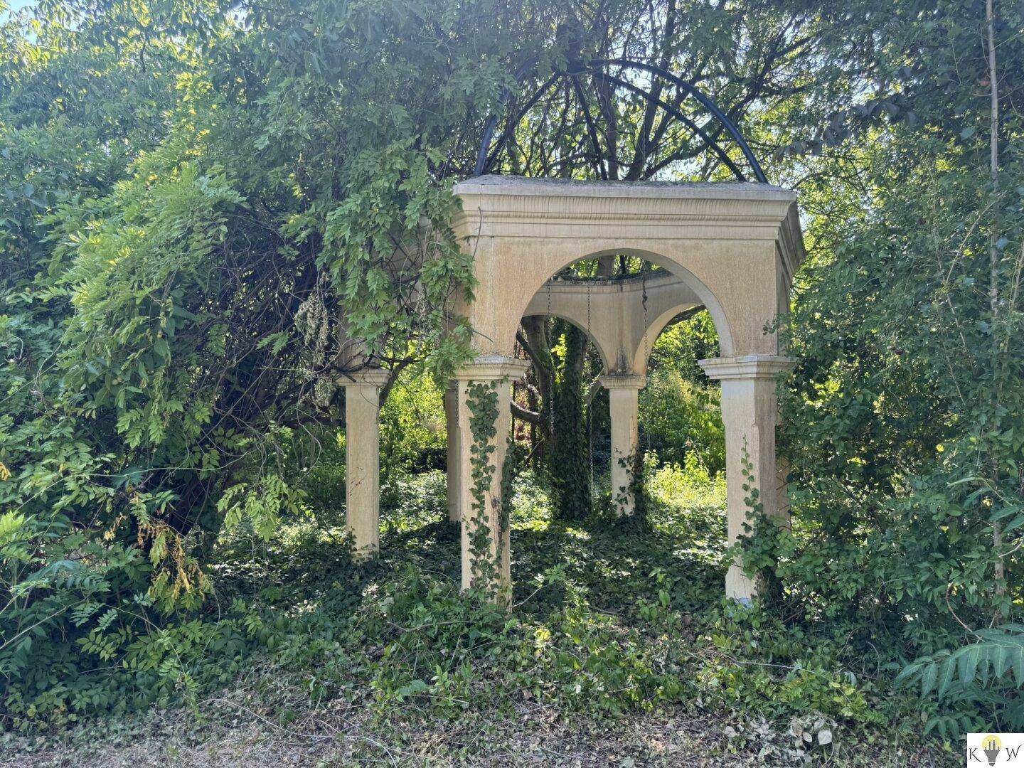 Eine alte, von Efeu überwucherte Steinlaube inmitten dichter Vegetation, die einen verwunschenen Charme versprüht.