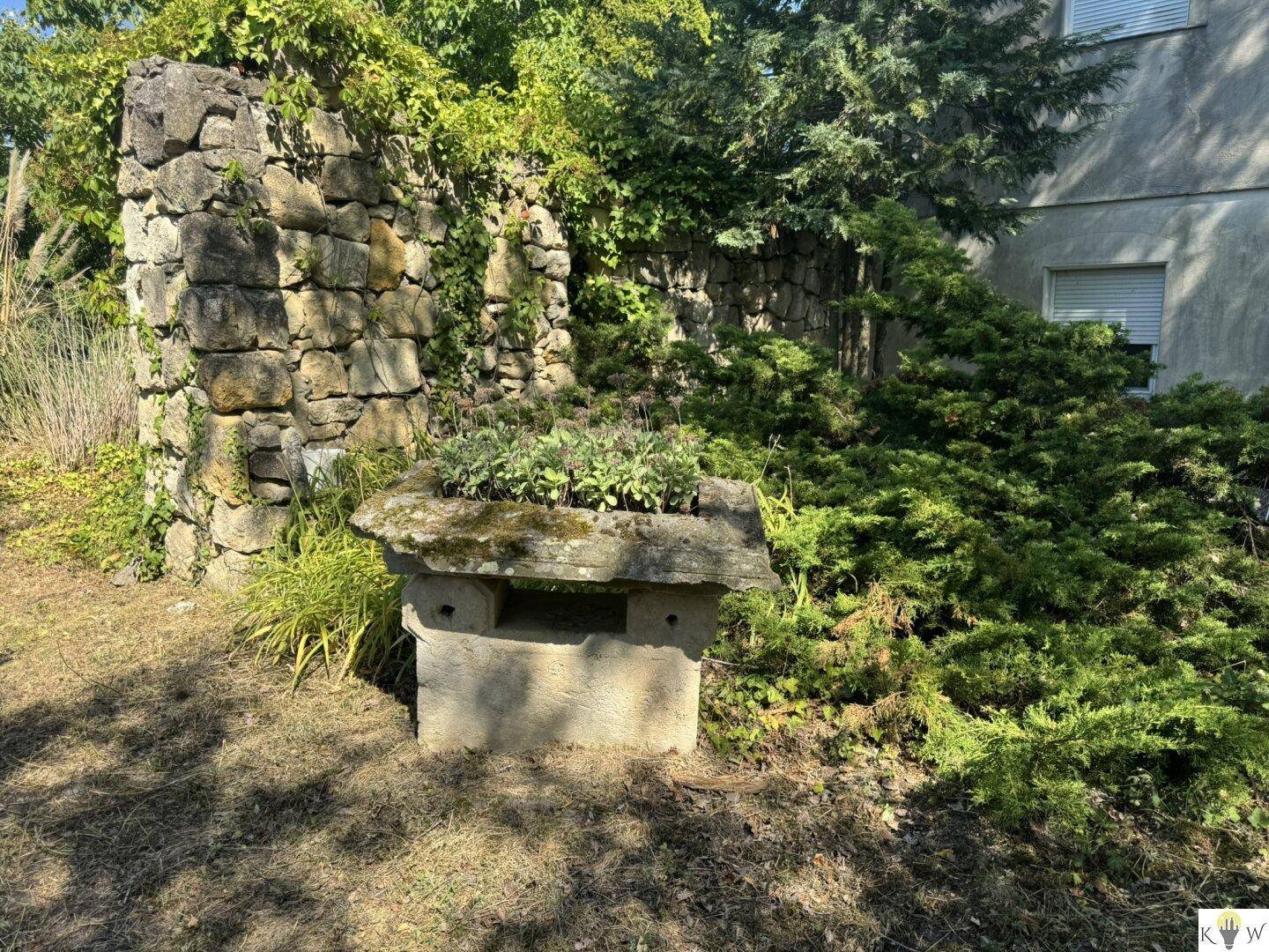 Rustikaler Gartenbereich mit alten Steinmauern und einem steinernen Pflanzgefäß, umgeben von Grün.