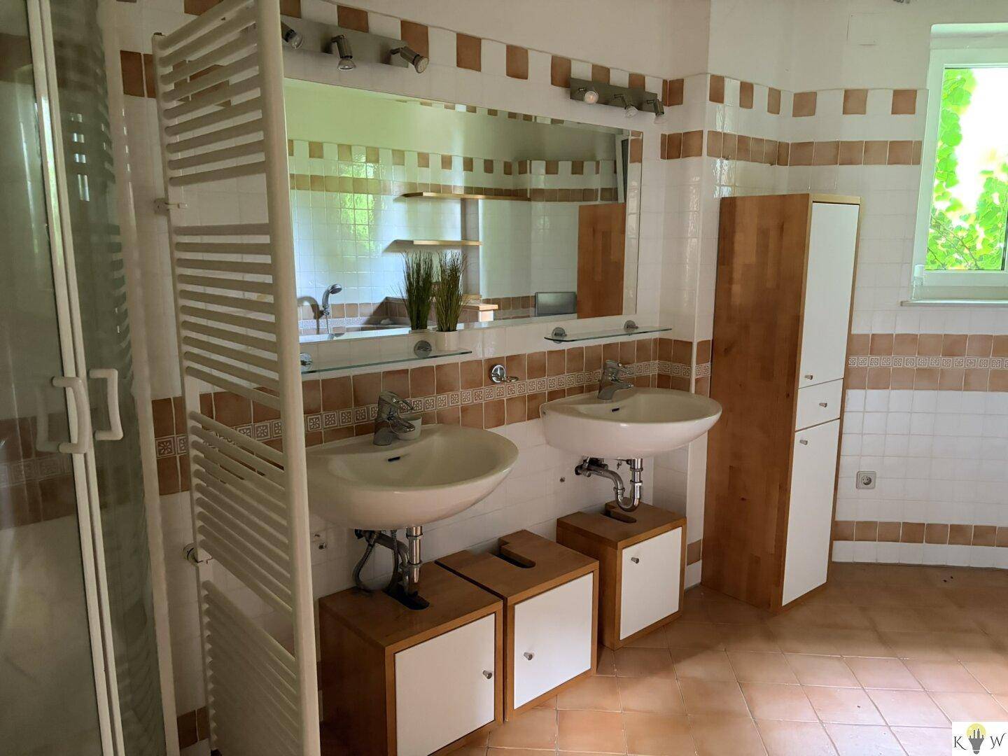 Badezimmer mit braun-weißen Fliesen, Doppelwaschbecken und großem Spiegel, ausgestattet mit einem Handtuchheizkörper.