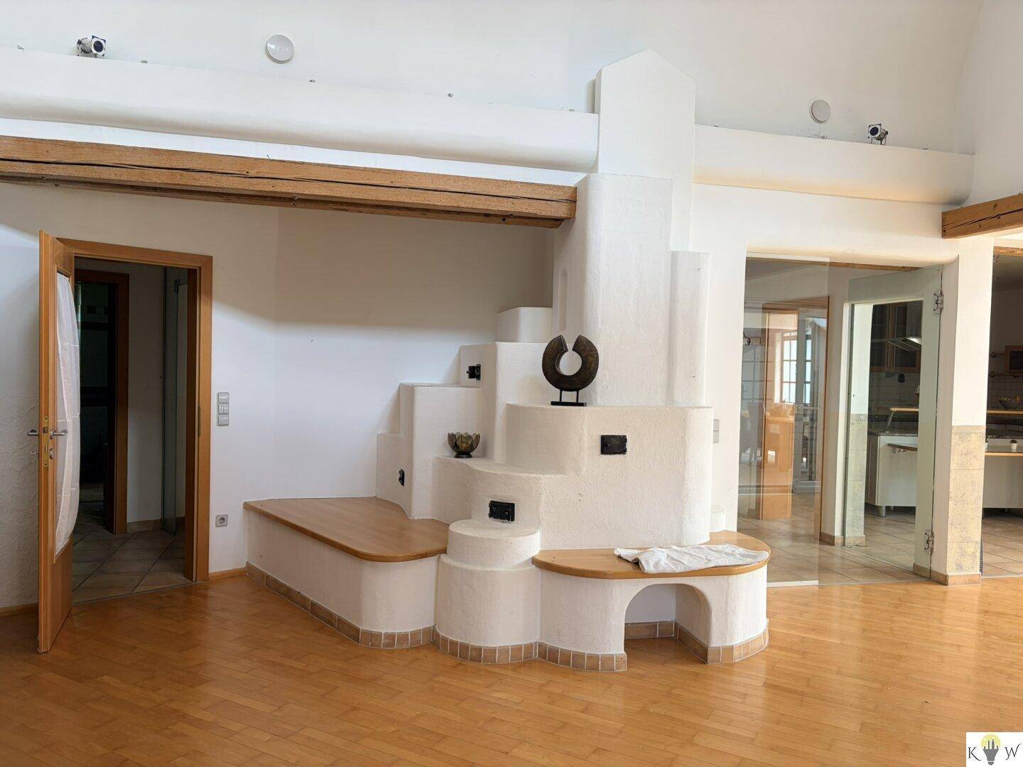Geräumiges Wohnzimmer mit Holzboden, sichtbaren Holzbalken und einem markanten, gestuften Kachelofen mit Sitzgelegenheiten.
