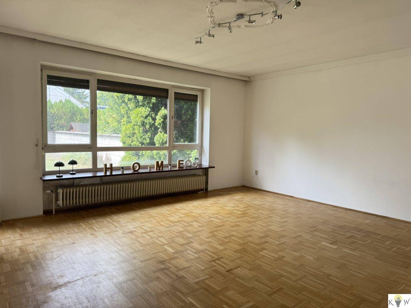 Großes Wohnzimmer mit Fischgrätparkett und breiter Fensterfront, die den Raum hell erscheinen lässt.