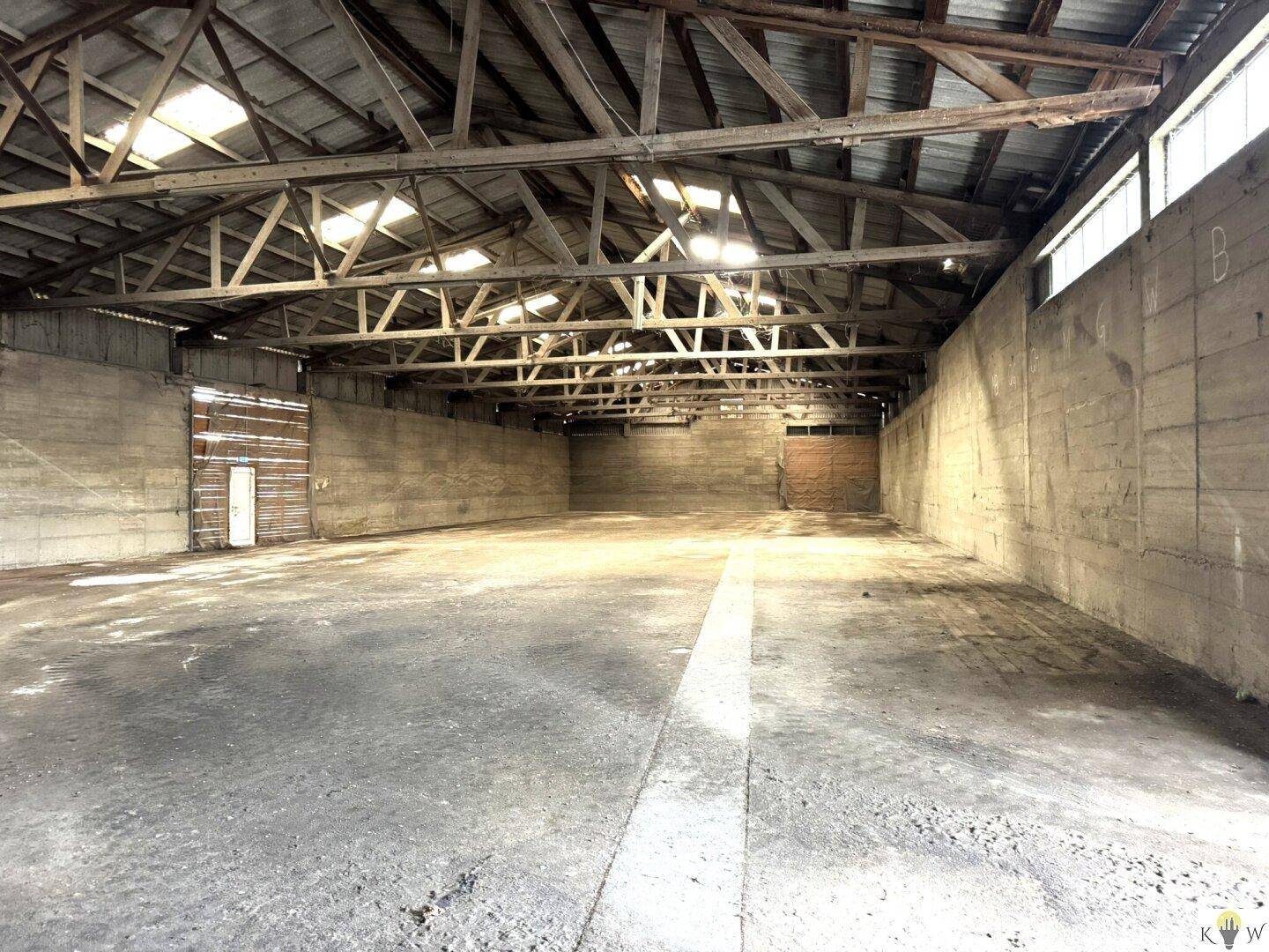 Geräumige, leere Lagerhalle mit Betonboden, hohen Betonwänden und sichtbarer Holz-Dachkonstruktion mit Oberlichtern.