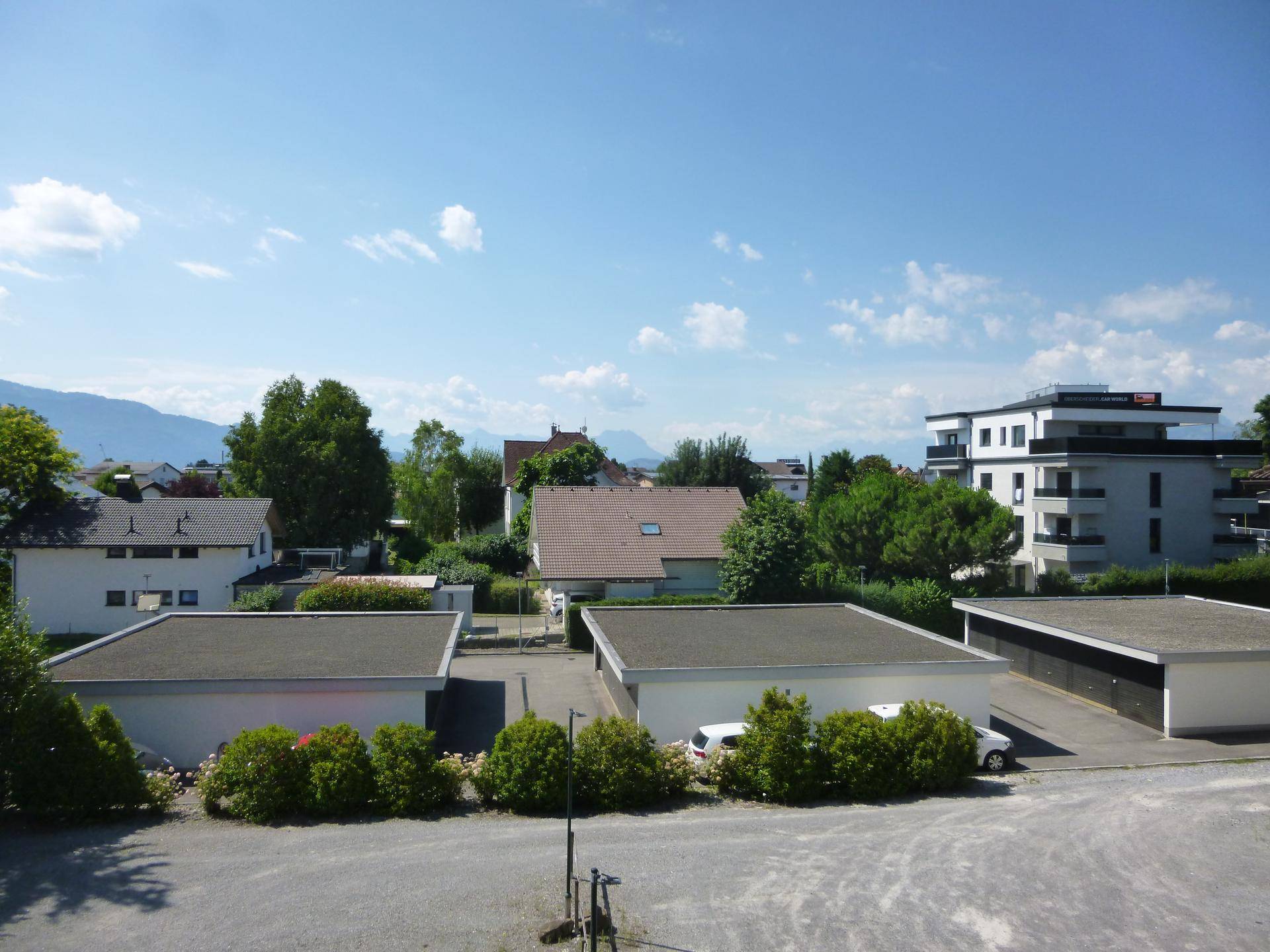 Weitblick über Dächer und umliegende Gebäude mit Bergen im Hintergrund unter blauem Himmel.