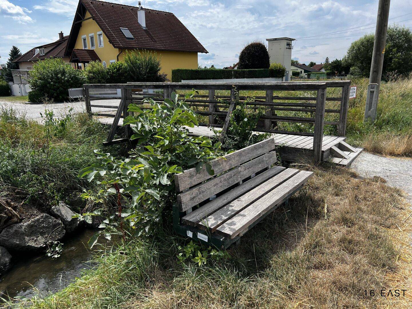 Holzbank am Bachufer mit einer Brücke und einem Wohnhaus im ländlichen Hintergrund.
