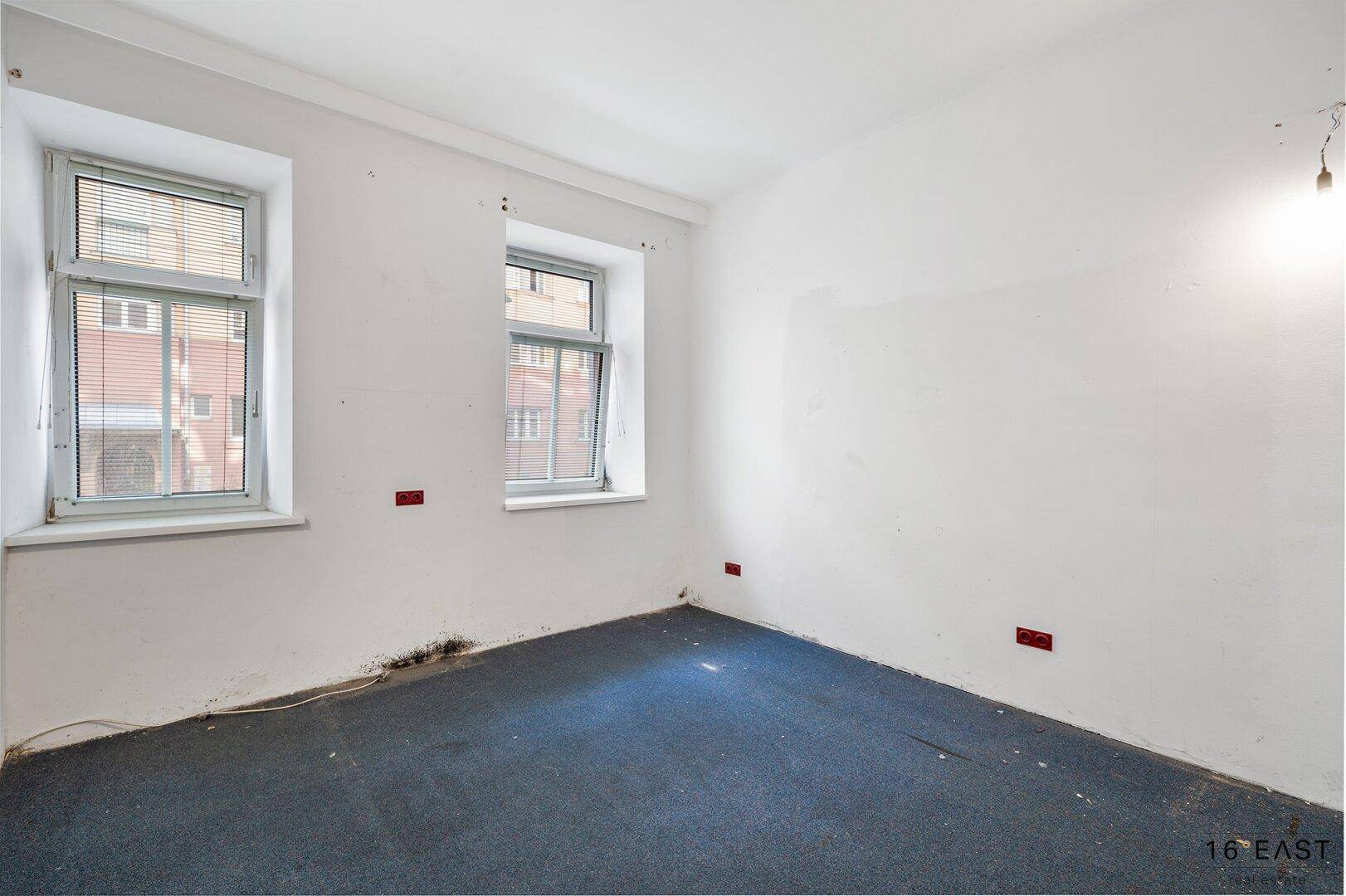 Leeres Schlafzimmer mit Teppichboden und zwei Fenstern, die Tageslicht hereinlassen.