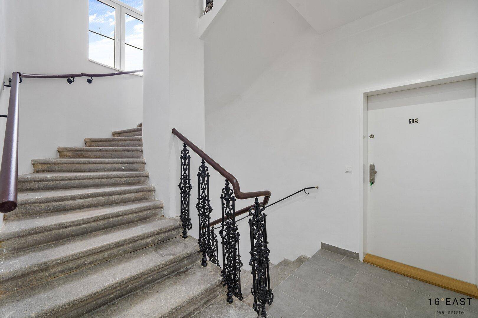 Treppenhaus mit Steinstufen, schmiedeeisernem Geländer und einem Fenster, das Tageslicht hereinlässt.