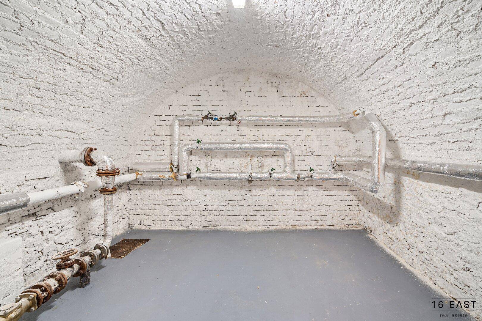 Kellerraum mit sichtbaren Rohren, gewölbter Decke und grauem Betonboden.