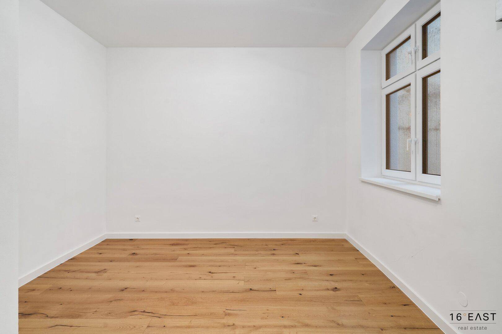 Helles, leeres Zimmer mit Holzboden und großem Fenster.