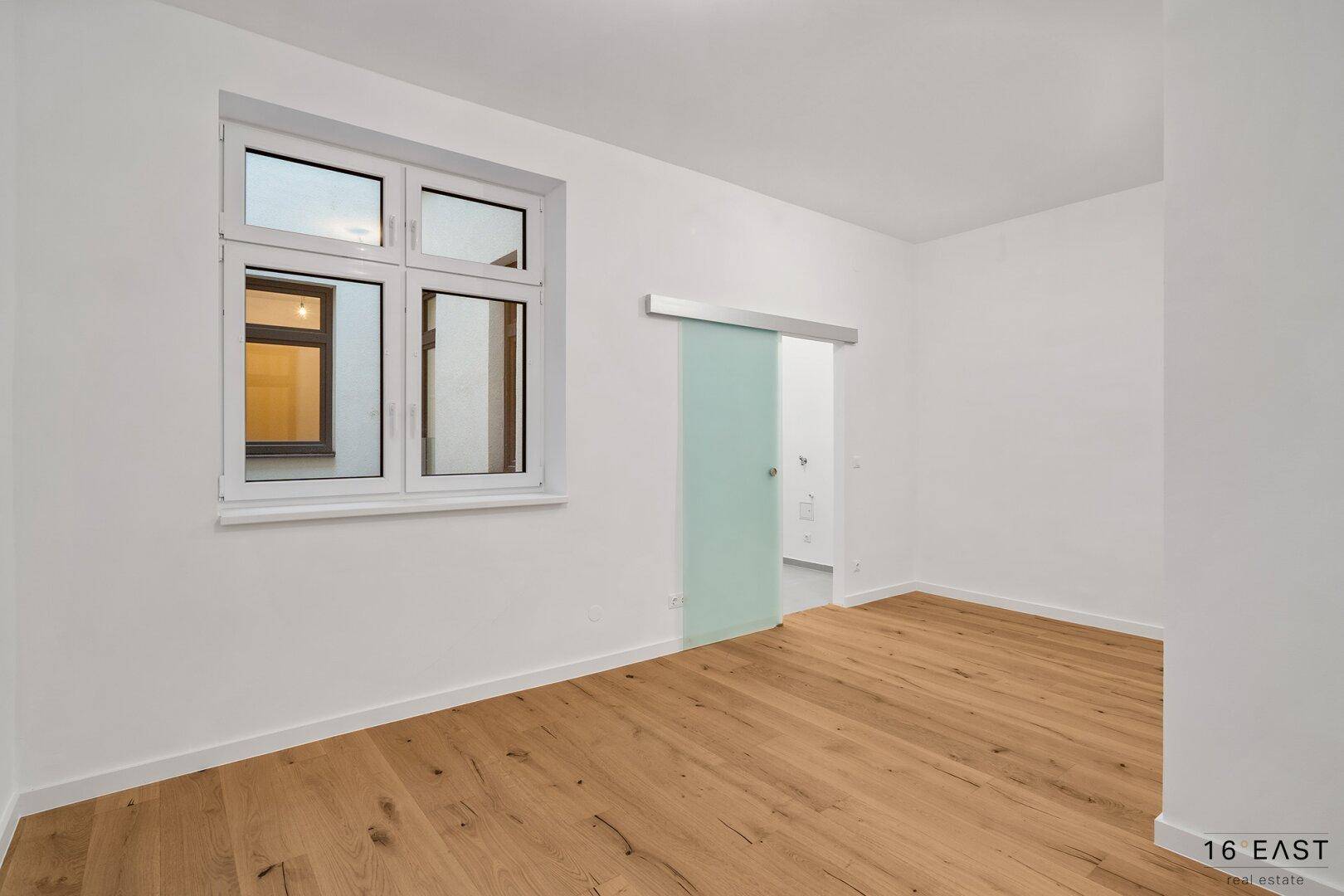 Leerer Raum mit Holzboden, Fenster und einer modernen Glasschiebetür.