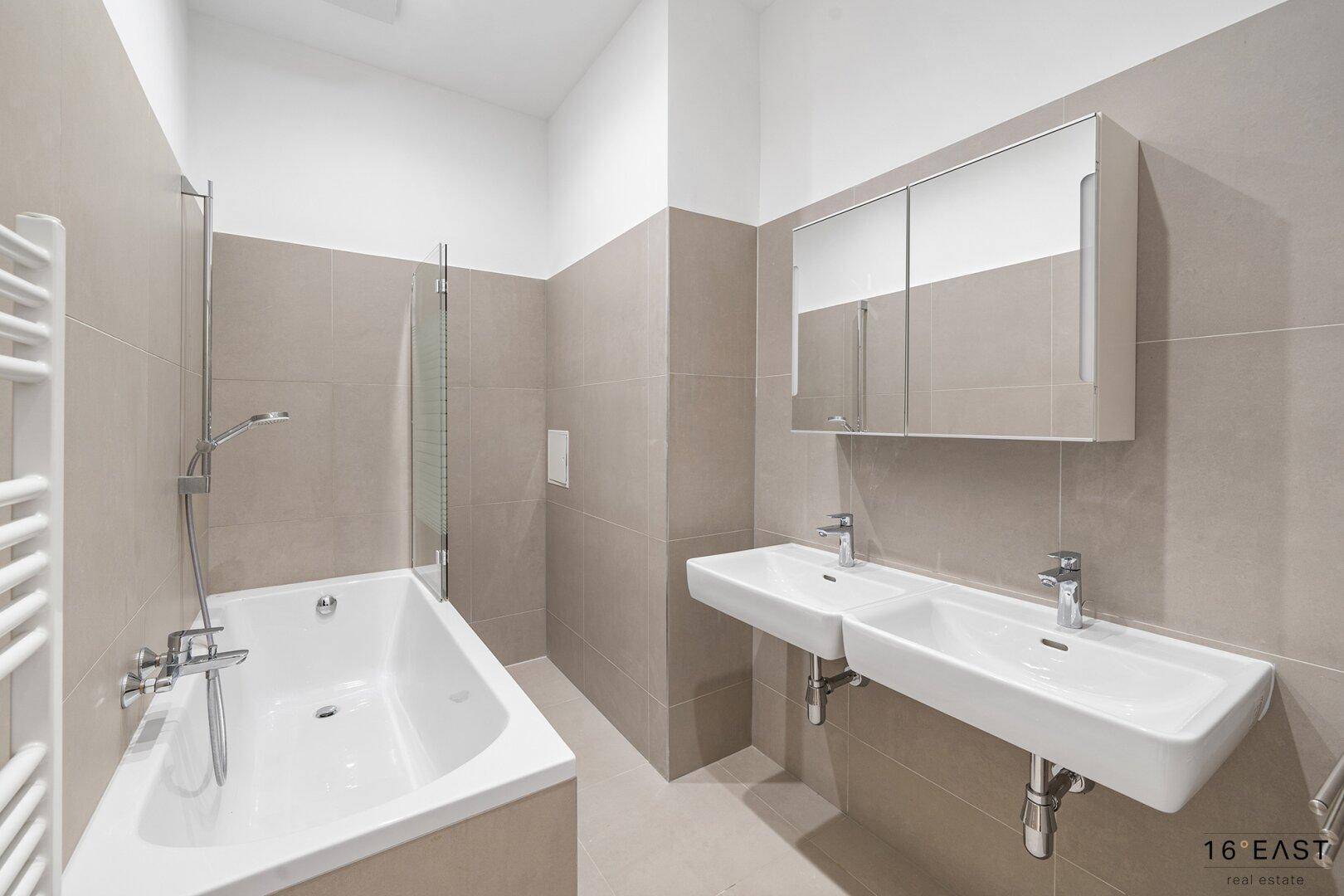 Modernes Badezimmer mit beigefarbenen Fliesen, Badewanne, Doppelwaschbecken und Spiegelschrank.