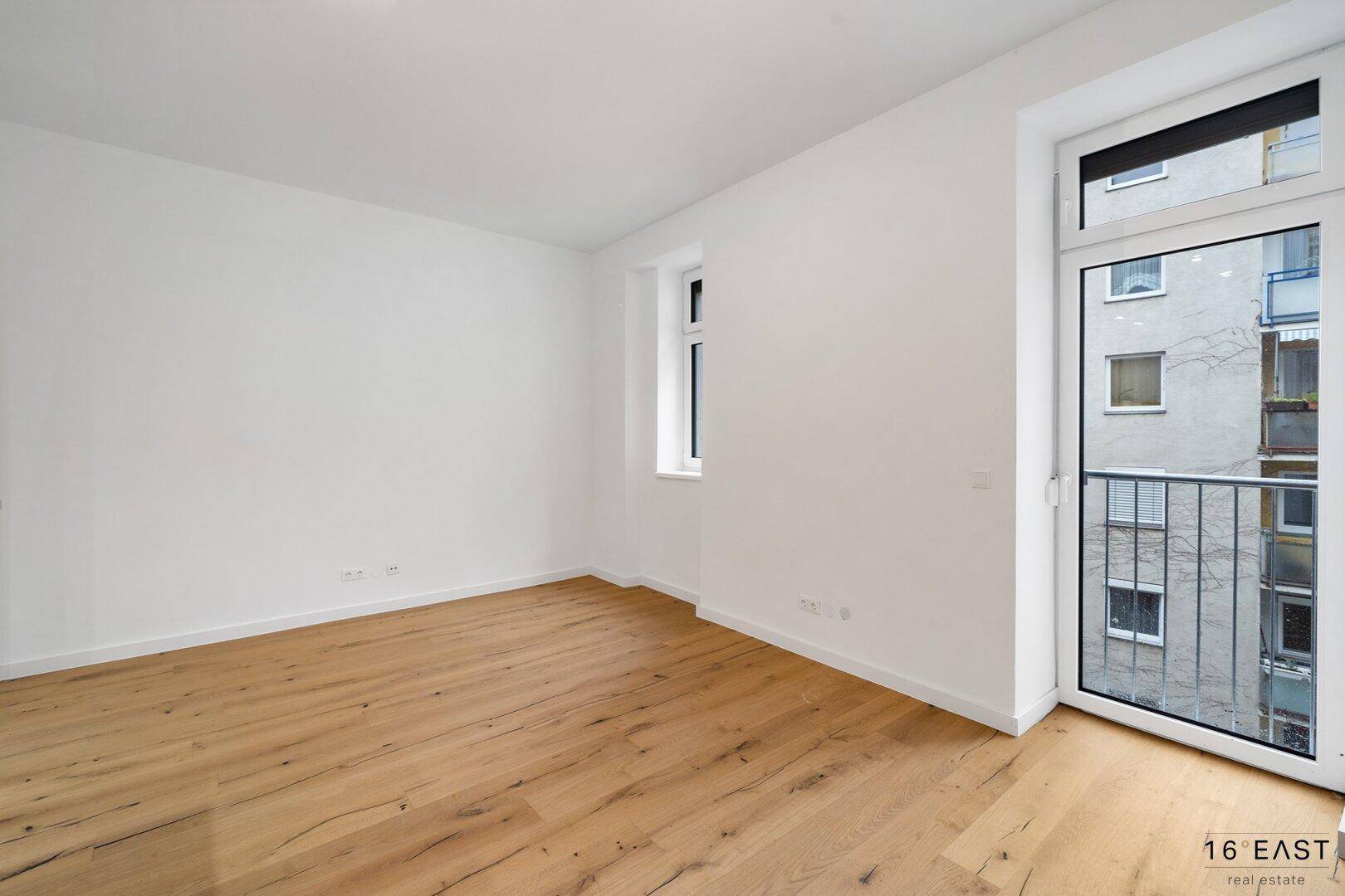 Leeres Zimmer mit hellem Holzboden, weißen Wänden und Zugang zu einem französischen Balkon.