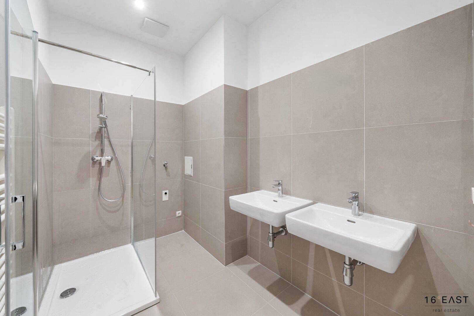 Modernes Badezimmer mit doppelten Waschbecken, großer Dusche und beigen Fliesen.
