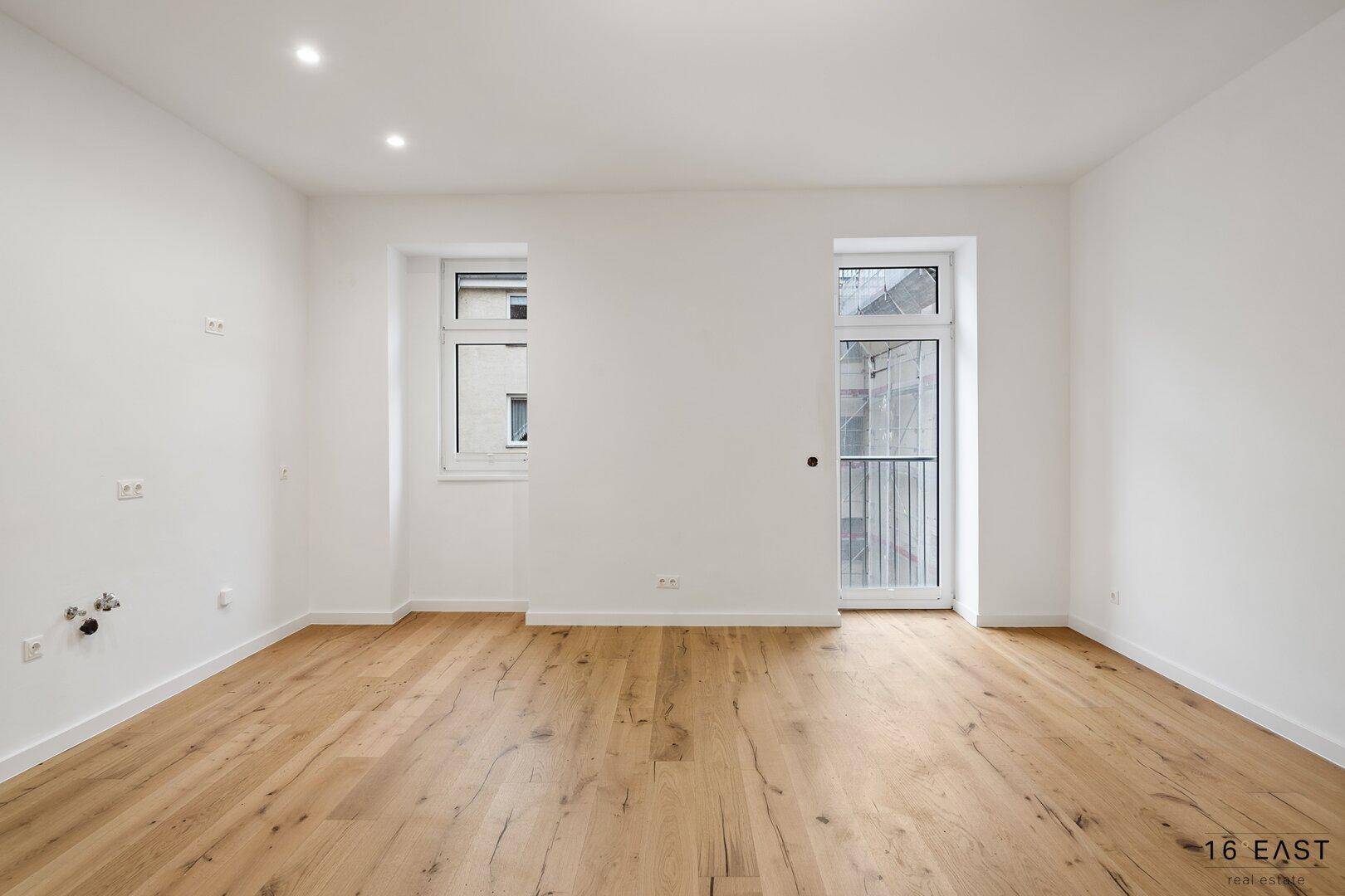 Geräumiges, unmöbliertes Zimmer mit hellem Holzboden und großen Fenstern.