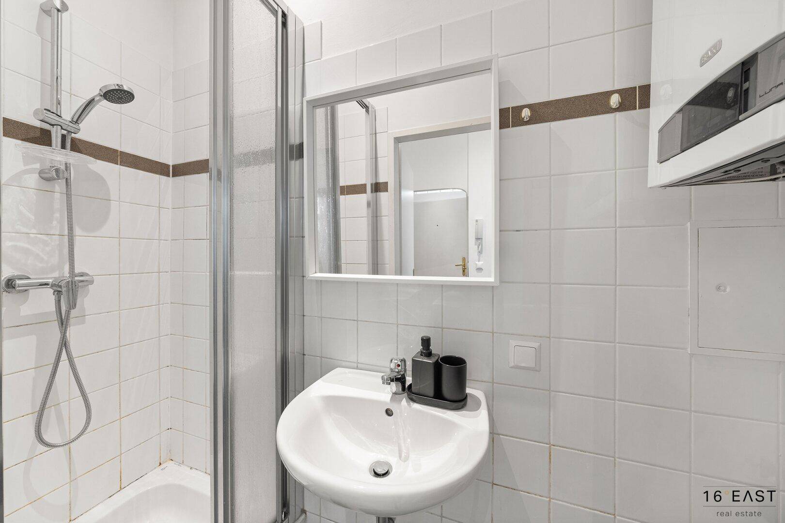 Kleines Badezimmer mit Duschkabine, Waschbecken, Spiegel und weiß gefliesten Wänden.