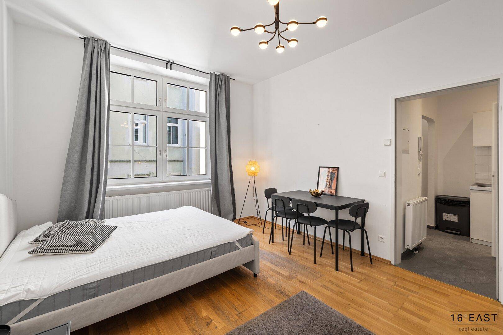 Studio-Apartment mit Schlafbereich, Esstisch und Zugang zu einer kleinen Küchenzeile durch einen Bogen.