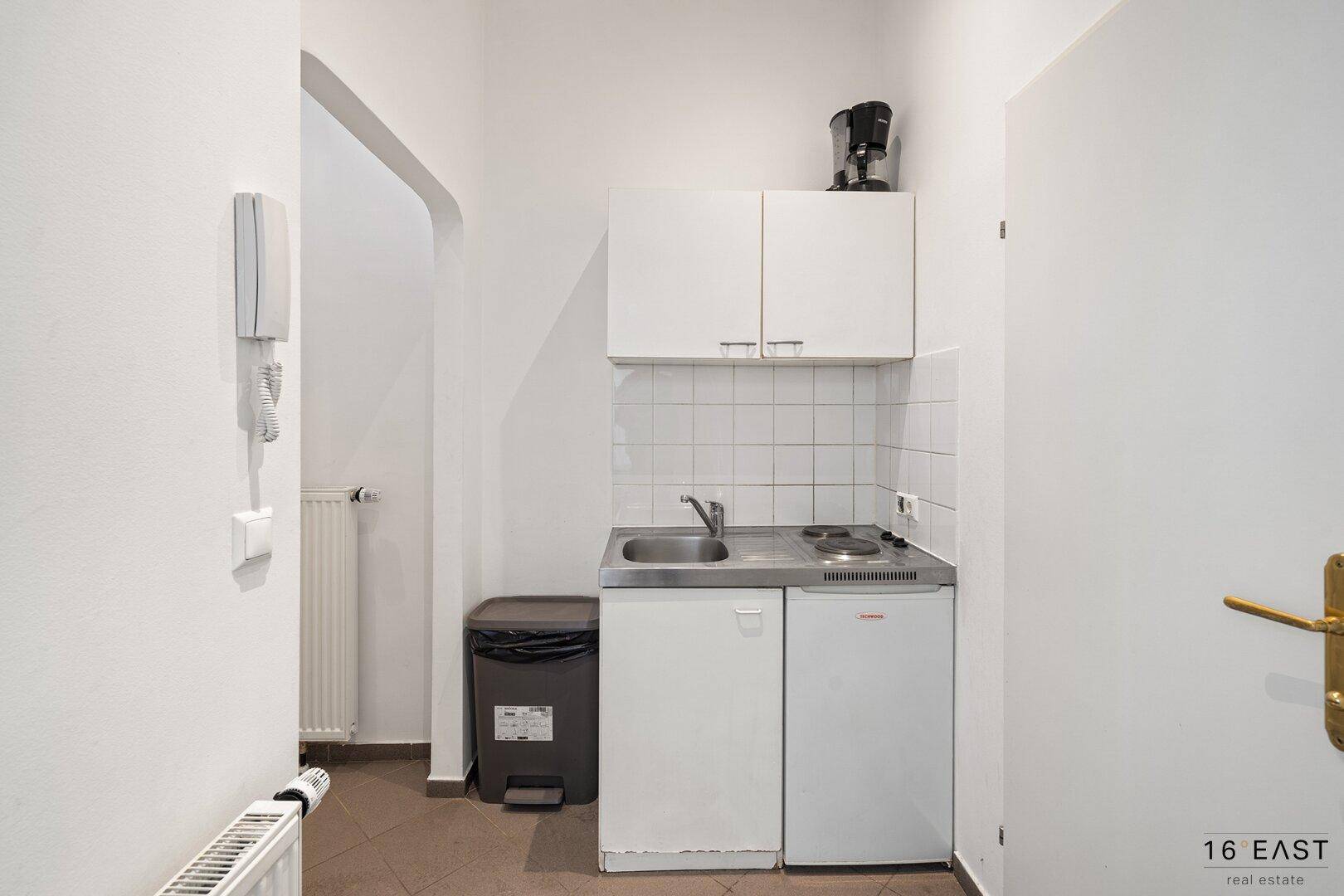 Kompakte Küchenzeile mit Spüle, Kochfeld, Kühlschrank und Kaffeemaschine in einem kleinen Raum.