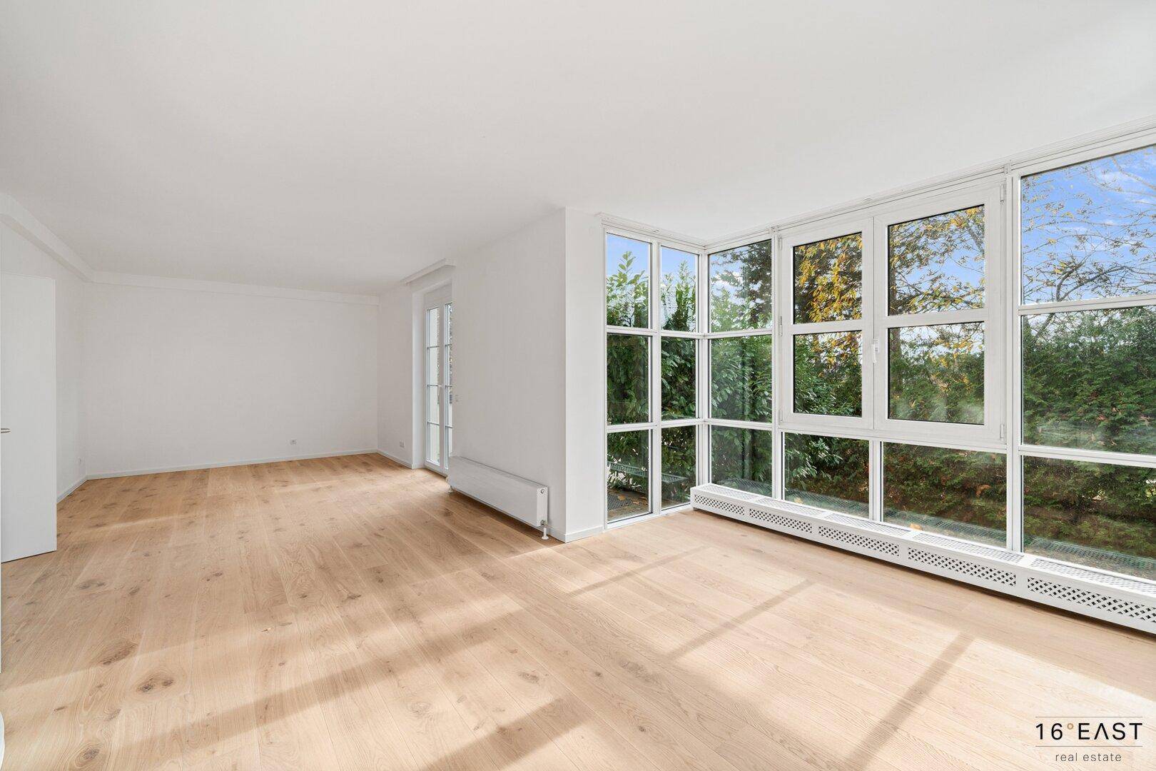 Unmöblierter Wohnbereich mit großen Fenstern, Holzboden und Heizkörpern unter den Fenstern.