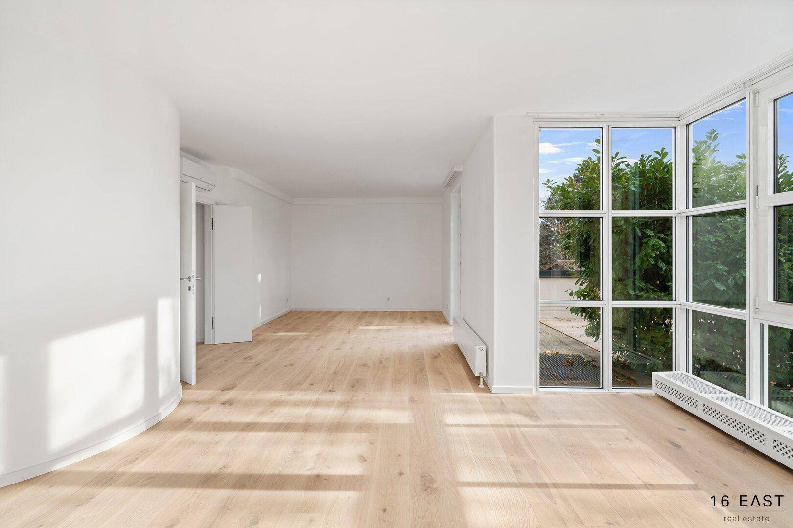 Leerer Wohnraum mit hellen Holzböden und großen Fenstern, die den Raum mit Licht füllen.