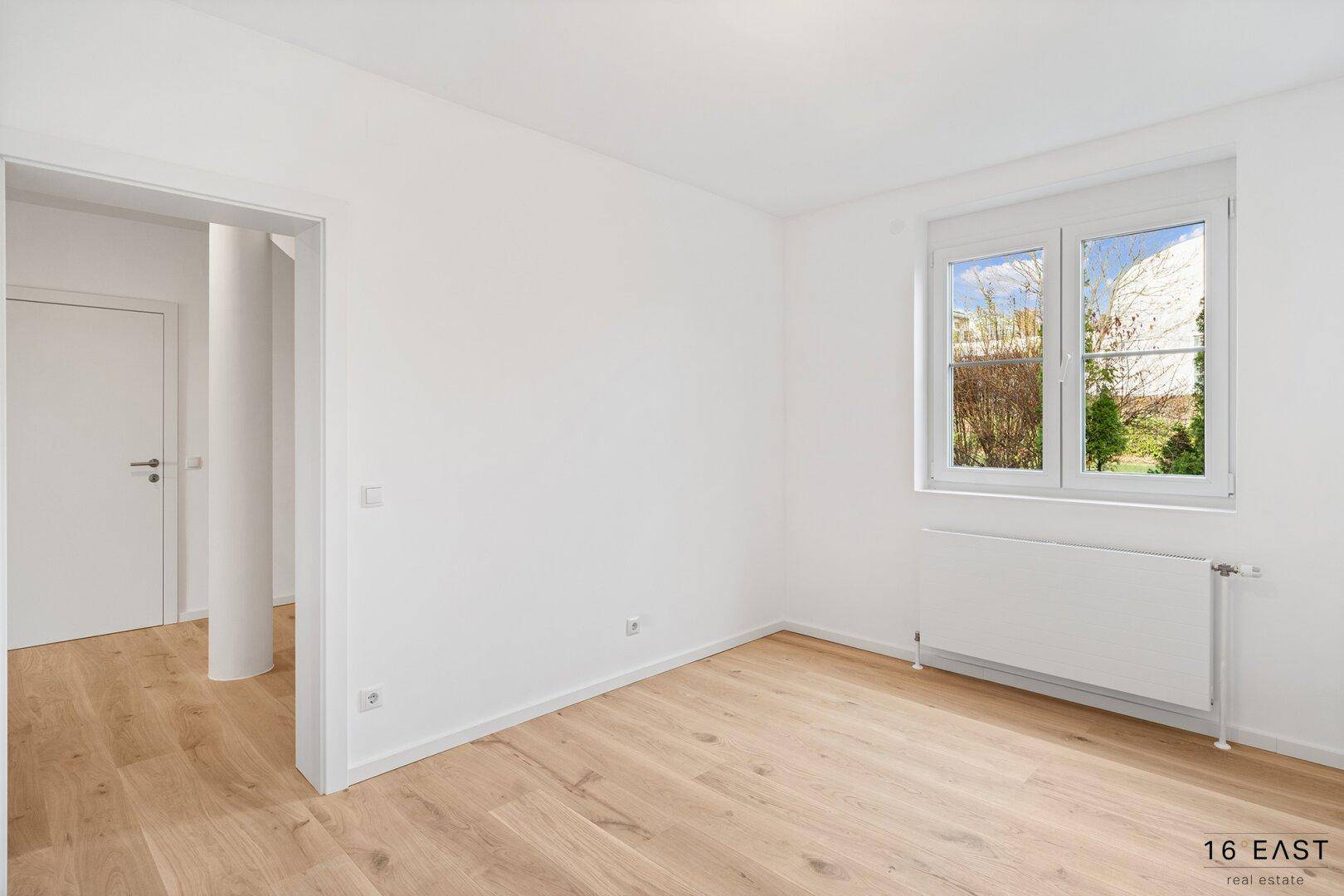 Leeres Schlafzimmer mit Holzboden und Fenster, das viel Tageslicht hereinlässt.