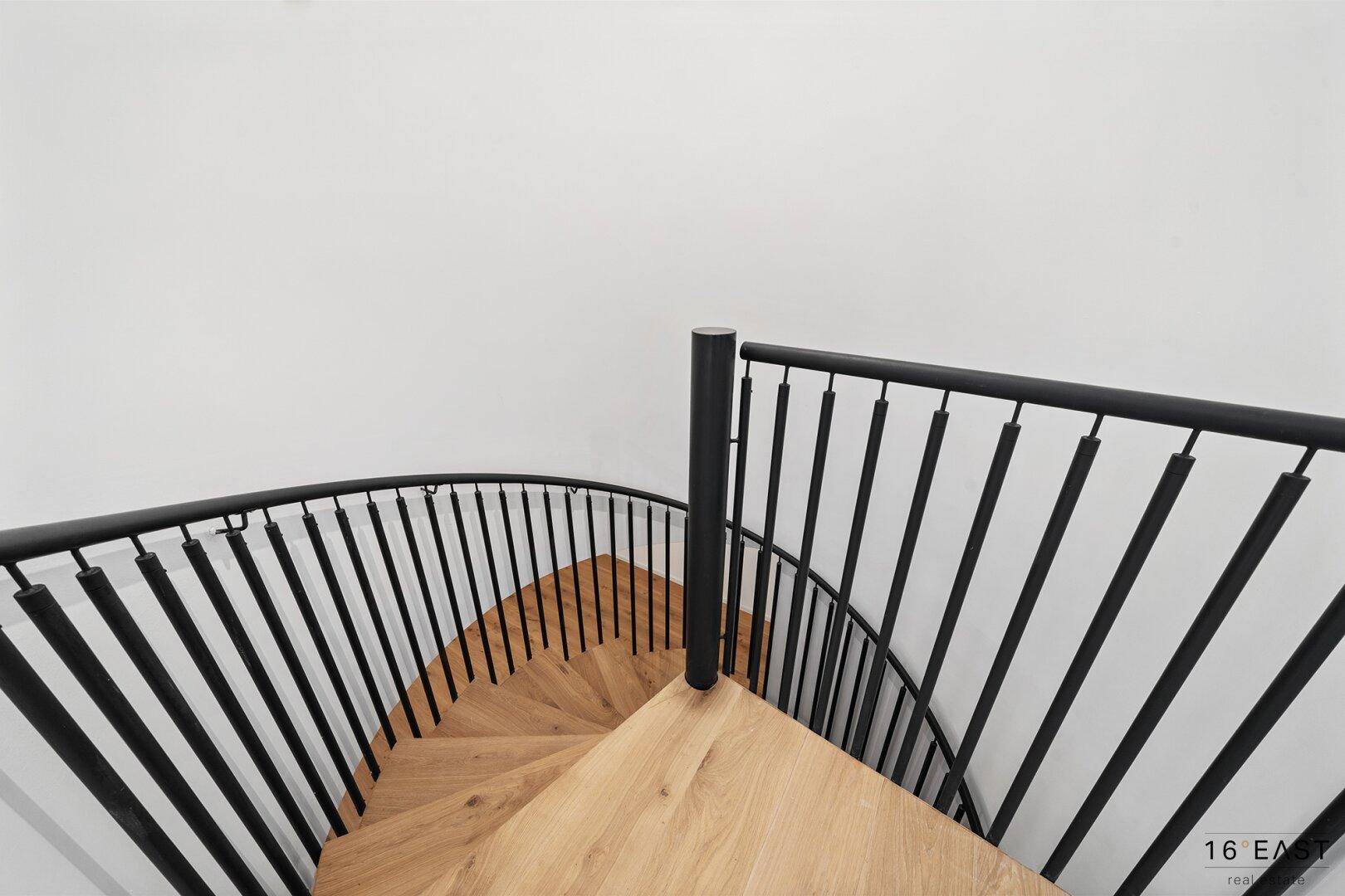 Moderne Wendeltreppe mit Holzstufen und schwarzem Metallgeländer, die verschiedene Ebenen verbindet.