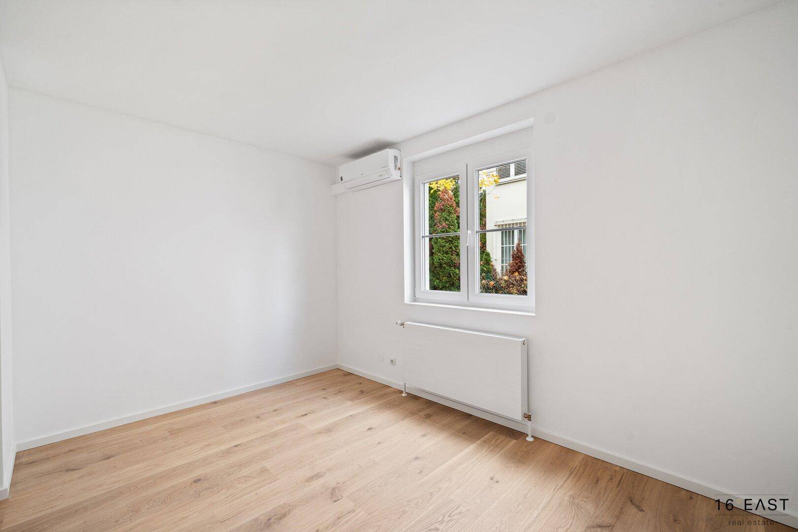 Helles Schlafzimmer mit Holzboden, Klimaanlage und Fenster mit Blick ins Grüne.