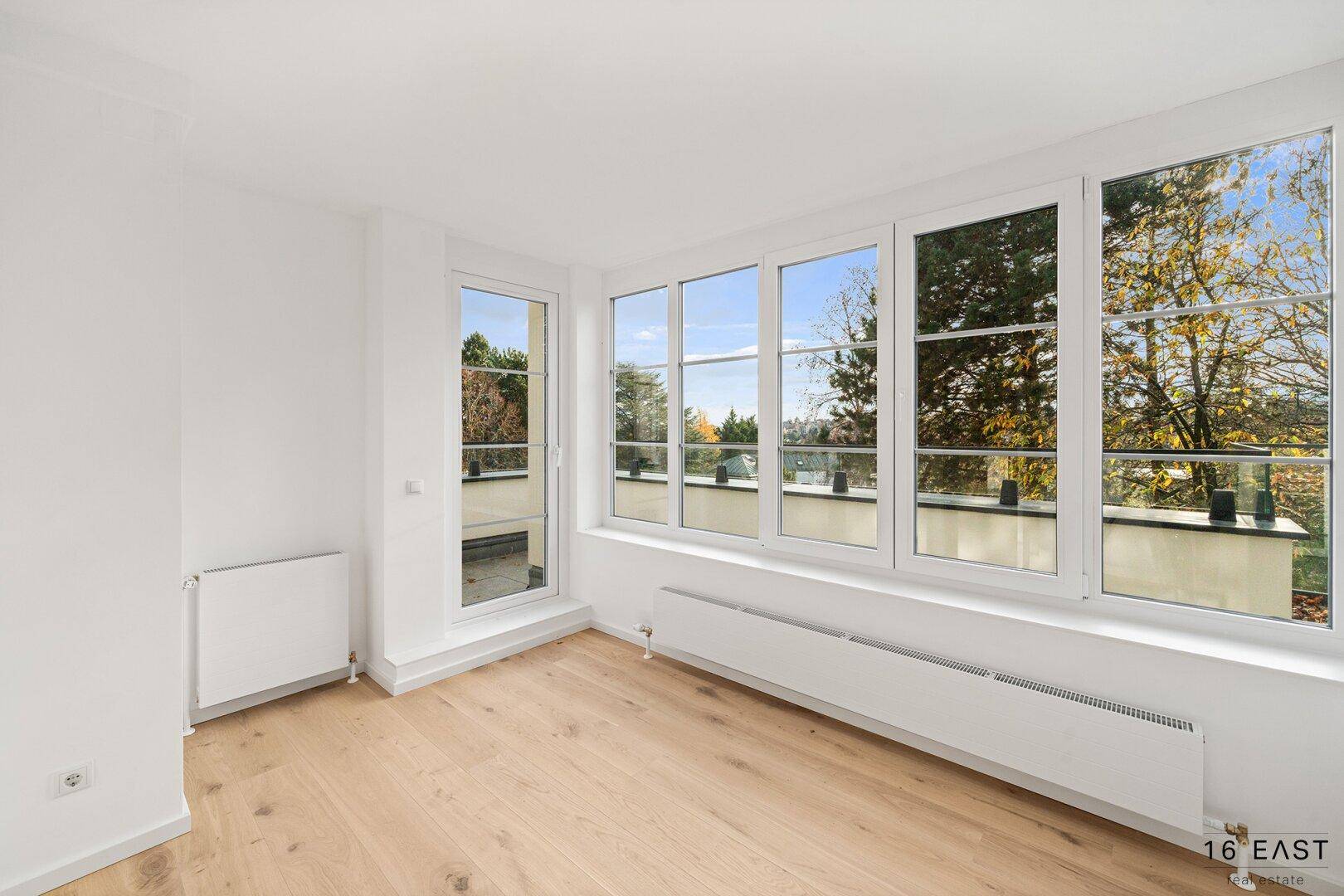 Helles Zimmer mit Holzboden und großen Fenstern, die viel Tageslicht hereinlassen und einen Blick ins Grüne bieten.