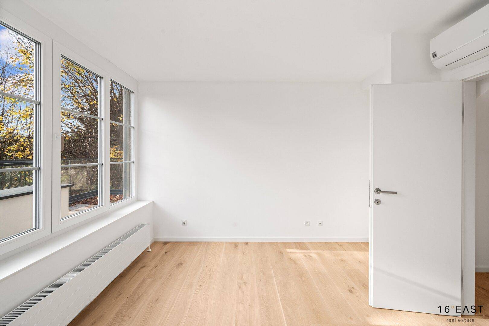 Unmöbliertes Zimmer mit Holzboden, großen Fenstern und Klimaanlage, bietet viel natürliches Licht.