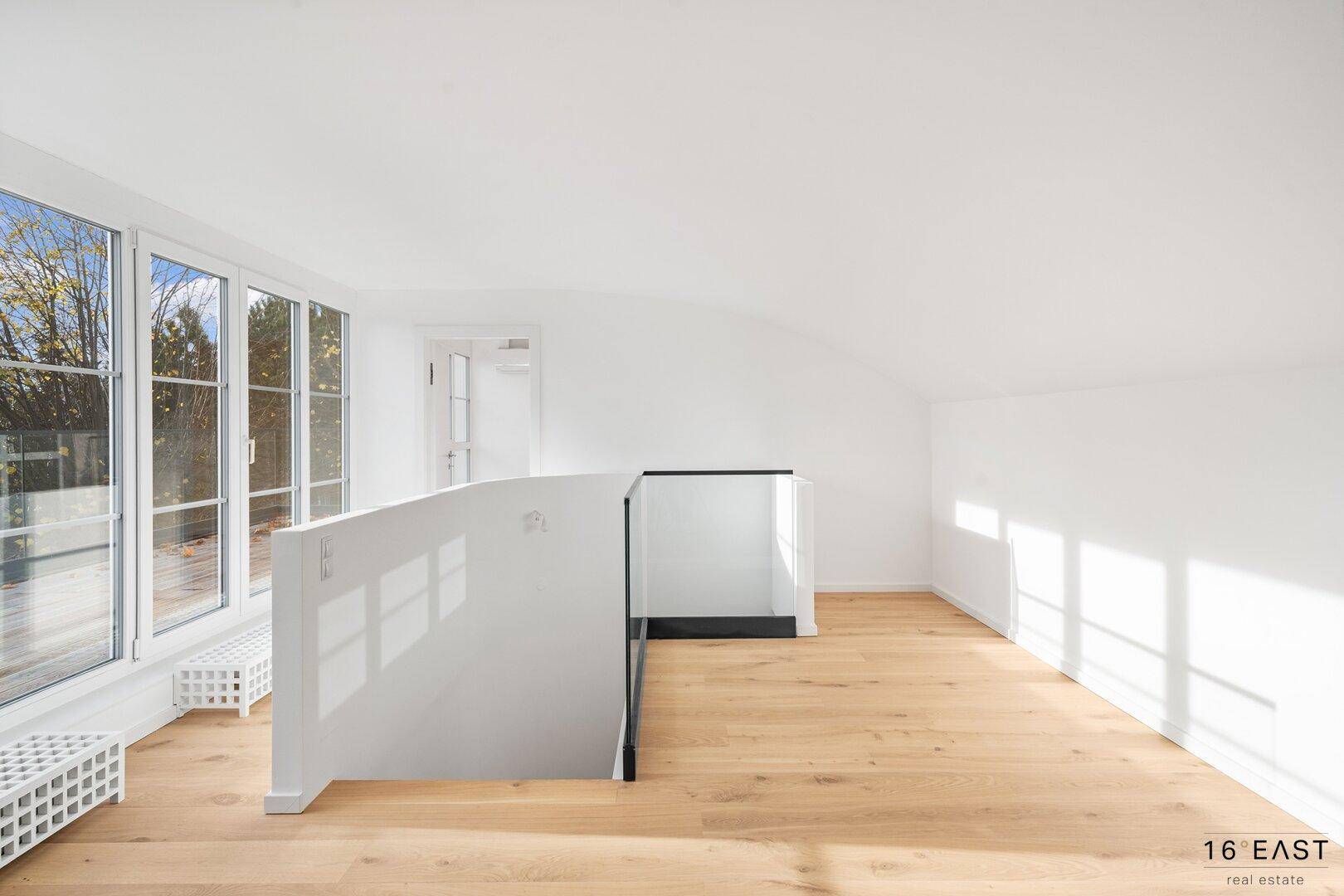 Offener Bereich mit Holzboden und moderner Treppe mit Glasgeländer, bietet eine helle und luftige Atmosphäre.