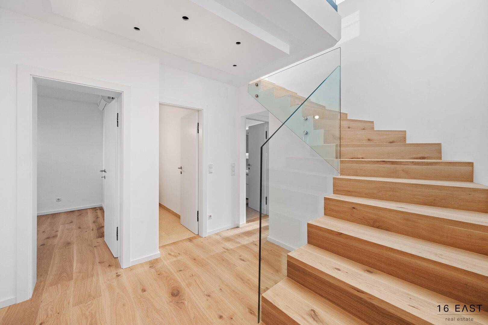 Heller Flur mit Holzboden und moderner Holztreppe mit Glasgeländer, führt zu weiteren Räumen.