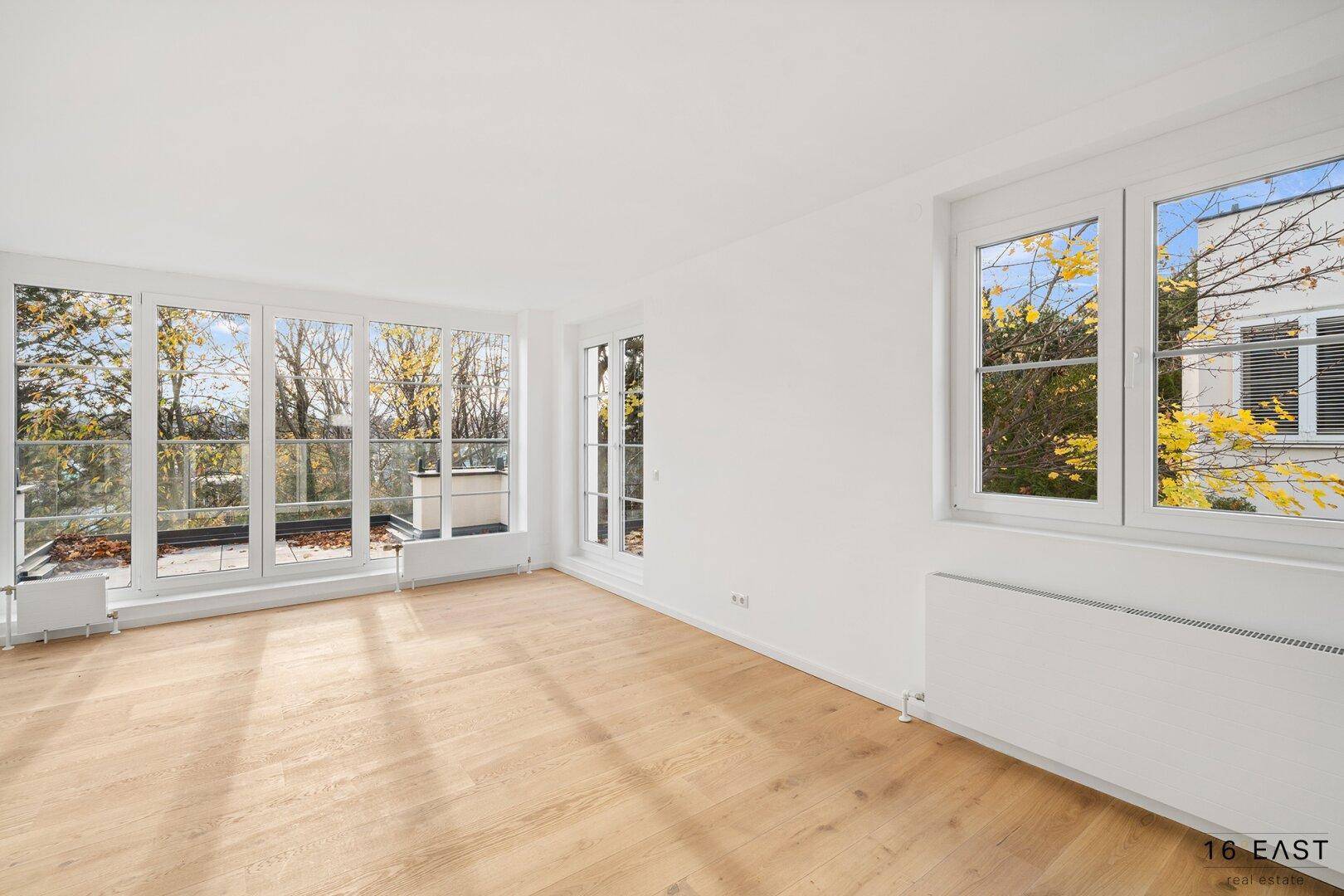 Geräumiger, unmöblierter Wohnbereich mit großen Fenstern, Holzboden und Zugang zur Terrasse, bietet viel Tageslicht.