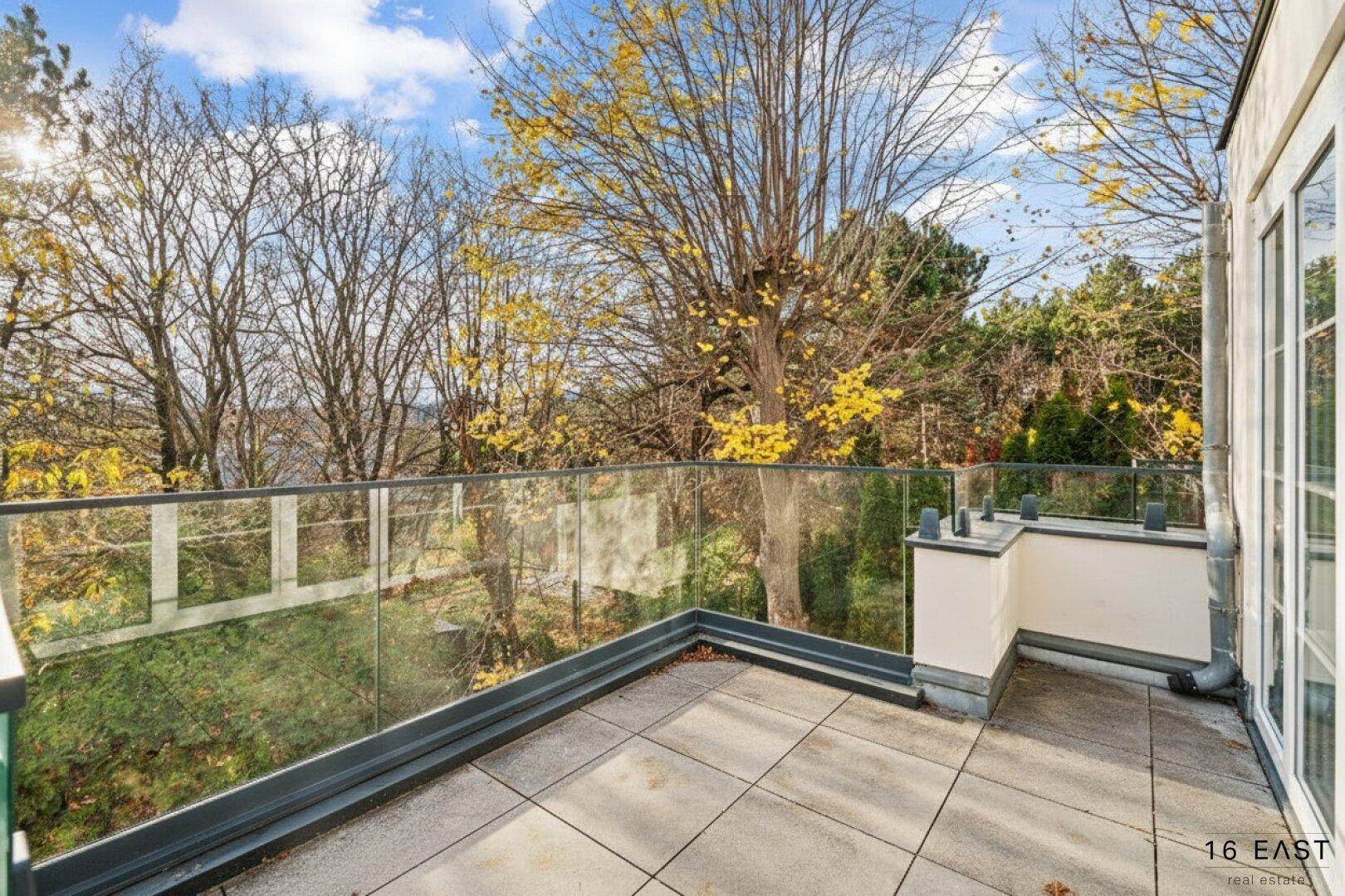 Geräumige Terrasse mit Fliesenboden und Glasgeländer, umgeben von Bäumen, bietet viel Platz im Freien.