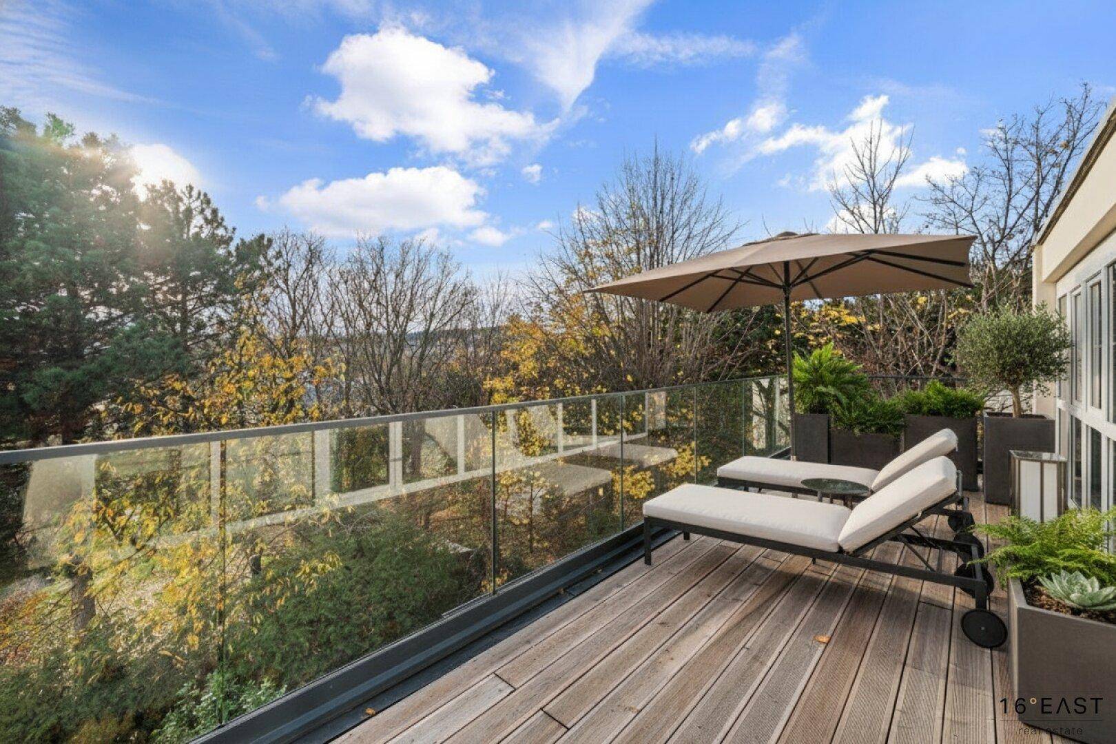 Große Holzterrasse mit Liegestühlen und Sonnenschirm, umgeben von Pflanzen, ideal zum Entspannen.