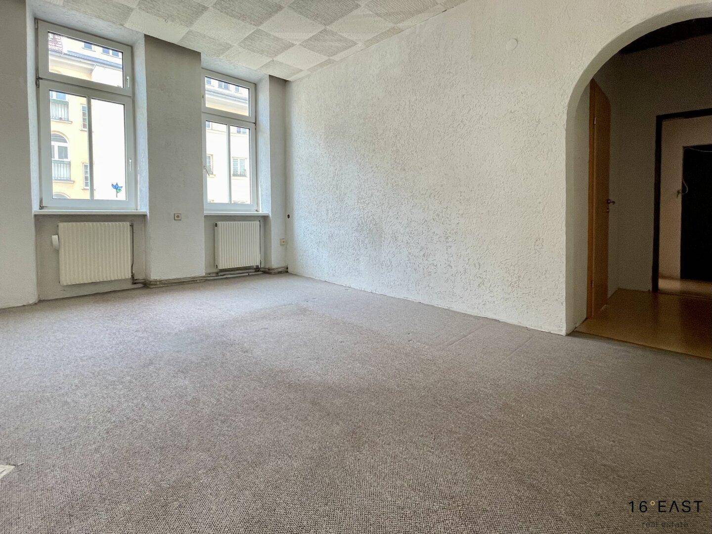 Geräumiges Wohnzimmer mit Teppichboden und zwei großen Fenstern, bereit zur Renovierung.