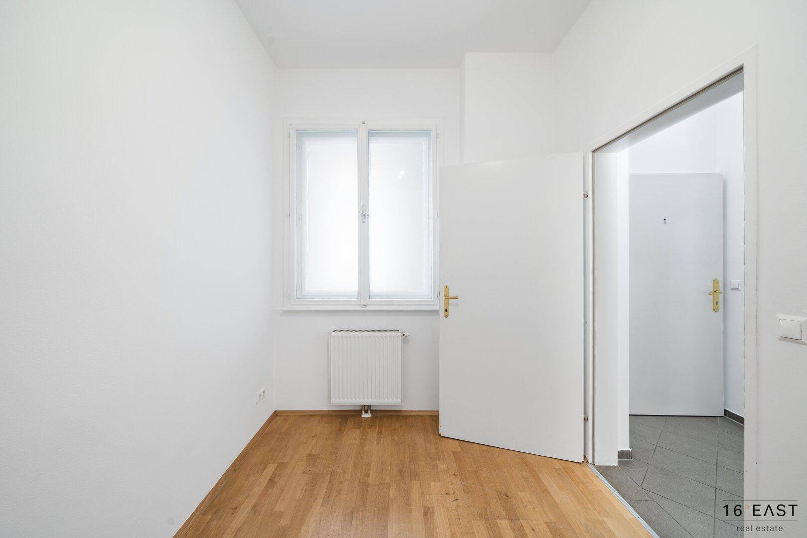 Kleines, helles Zimmer mit Holzfußboden und einem Fenster, das viel Tageslicht hereinlässt.