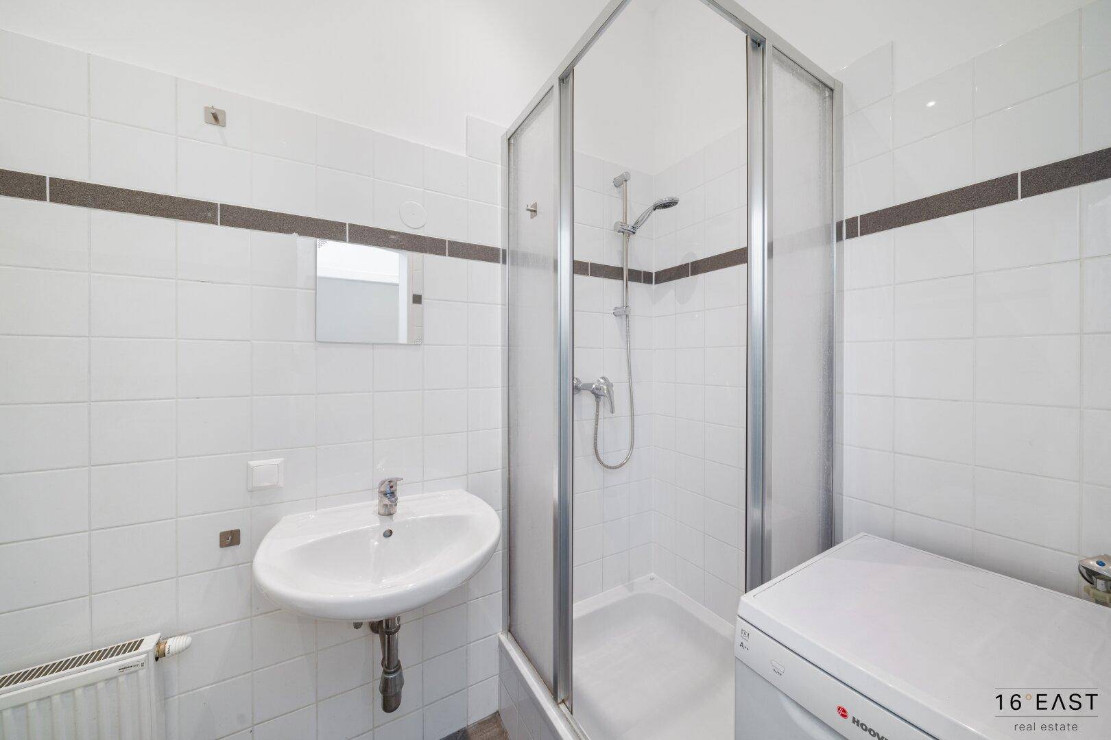 Gefliestes Badezimmer mit Duschkabine, Waschbecken und Platz für eine Waschmaschine.