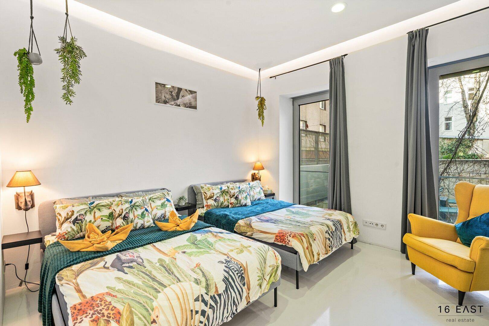 Helles Schlafzimmer mit zwei Betten, farbenfroher Bettwäsche und großen Fenstern.