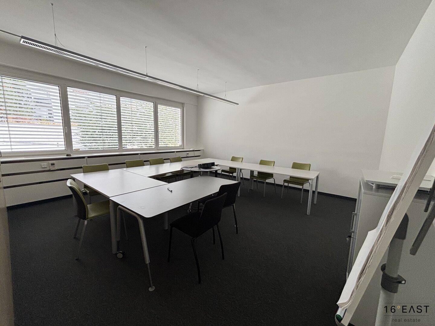 Großer Konferenzraum mit langen Fenstern, Teppichboden und mehreren Tischen für Besprechungen.