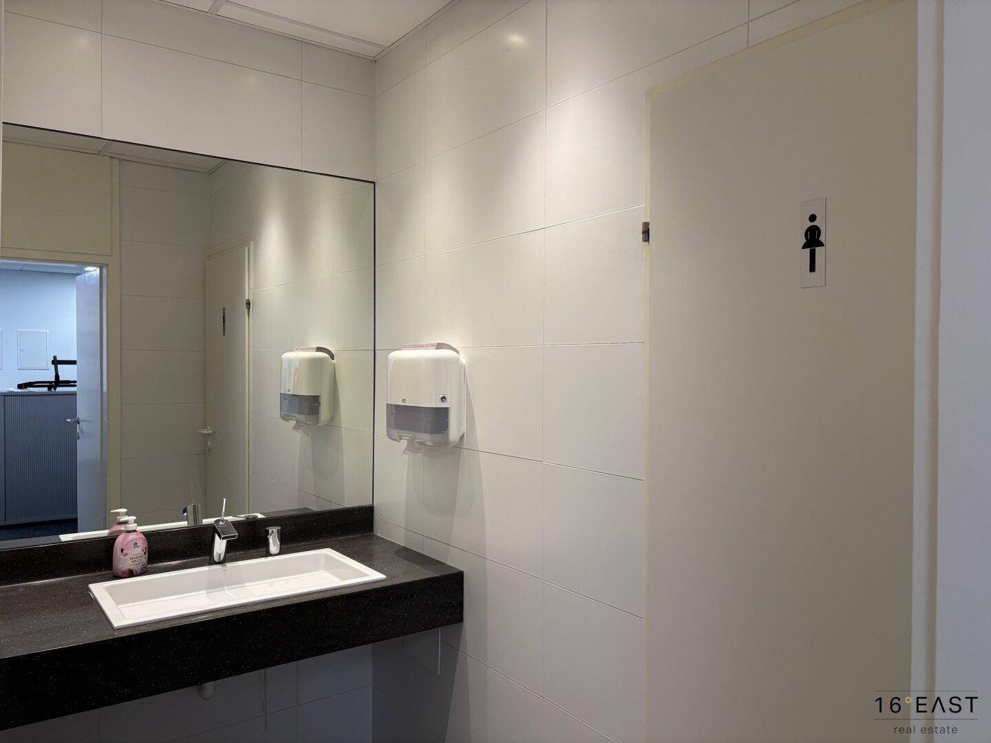 Saubere Toilette mit großem Spiegel, Waschbecken und weißen Fliesen, gut gepflegt.