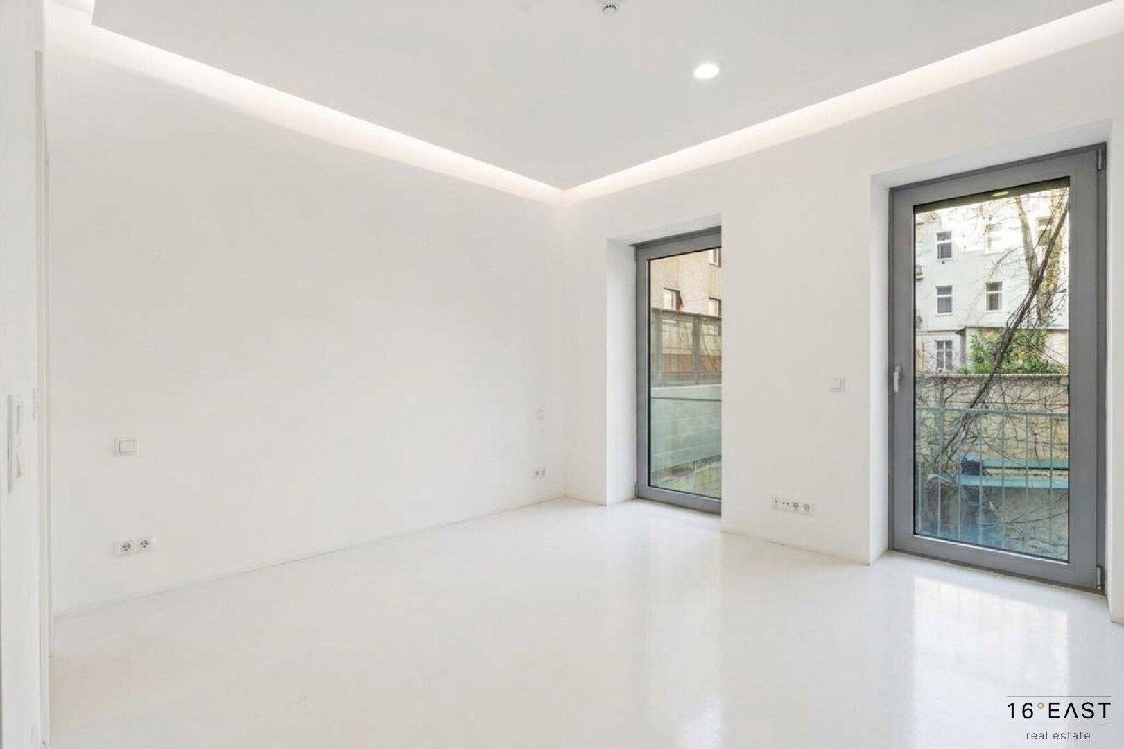Leerer, heller Raum mit großen Fenstertüren und indirekter Deckenbeleuchtung.