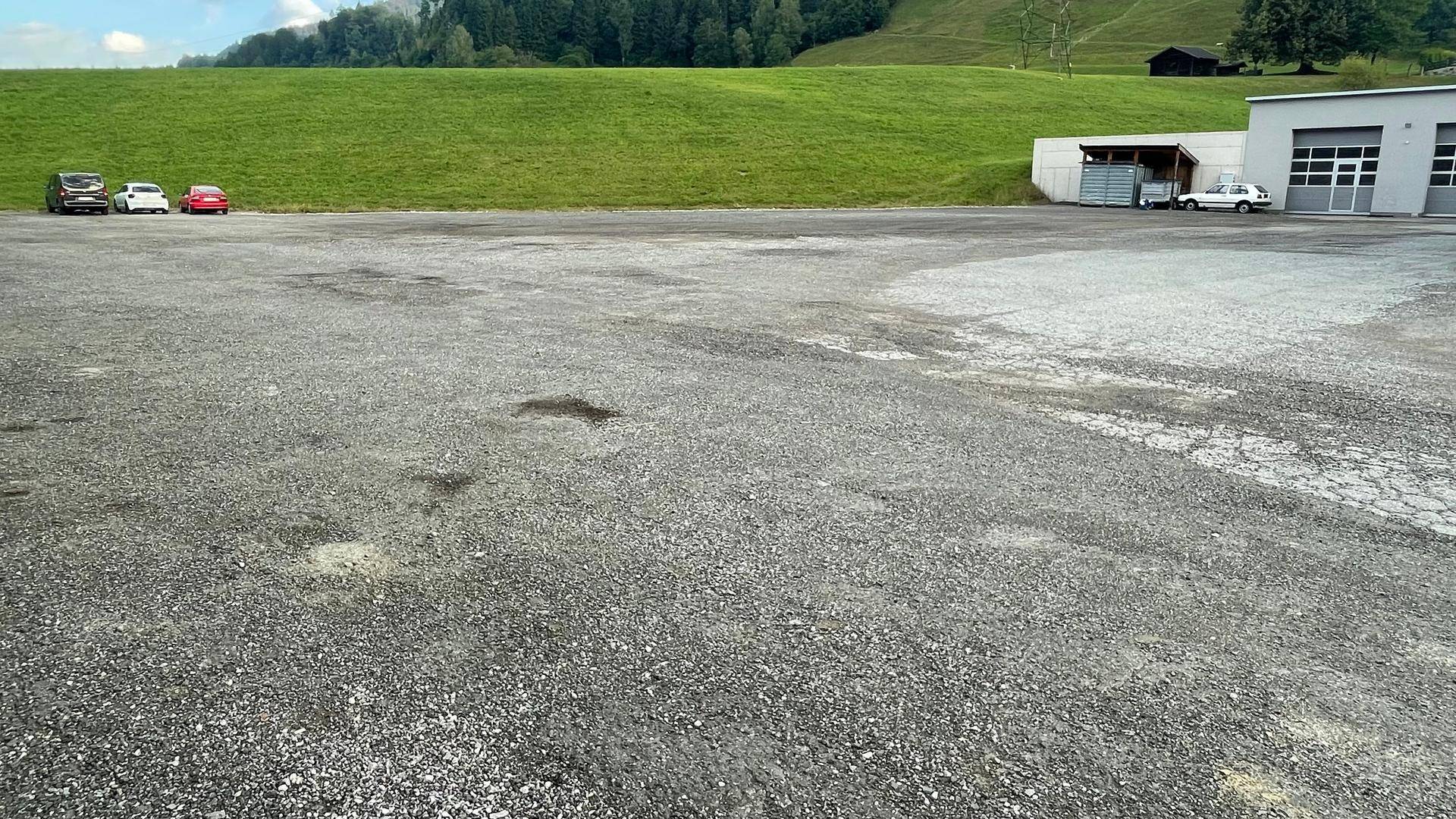 Große Freifläche aus Schotter und Asphalt vor einem Gebäude mit Garagen, umgeben von sanften grünen Hügeln und Bäumen.