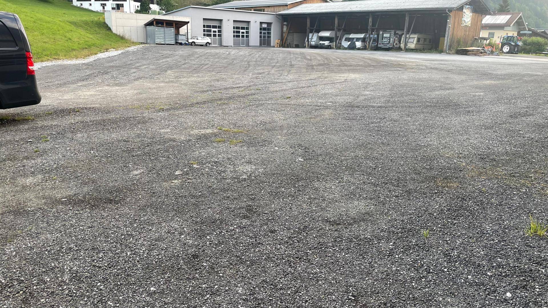 Weitläufiger Schotterparkplatz vor einem Gebäude mit Garagen und einem überdachten Bereich für Fahrzeuge, umgeben von Natur.