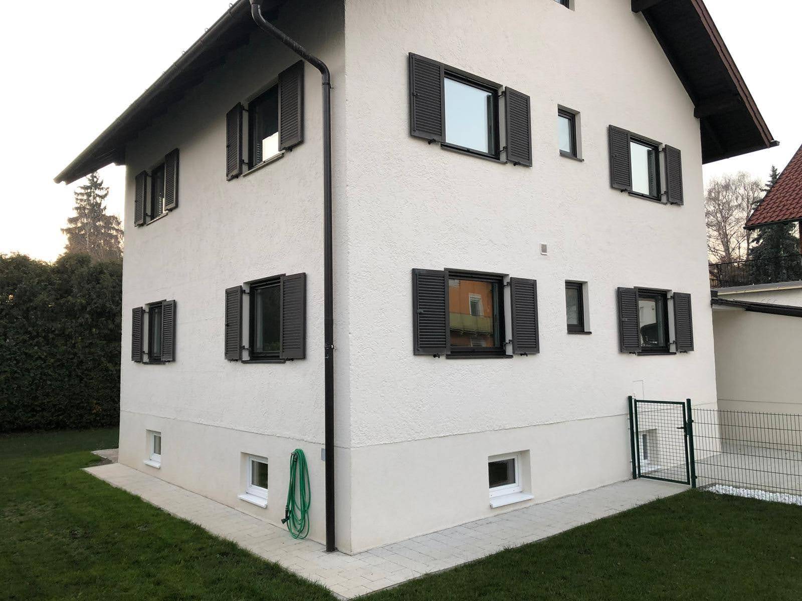 Seitenansicht des Hauses mit weißer Fassade, dunklen Fensterläden und grünem Rasen.