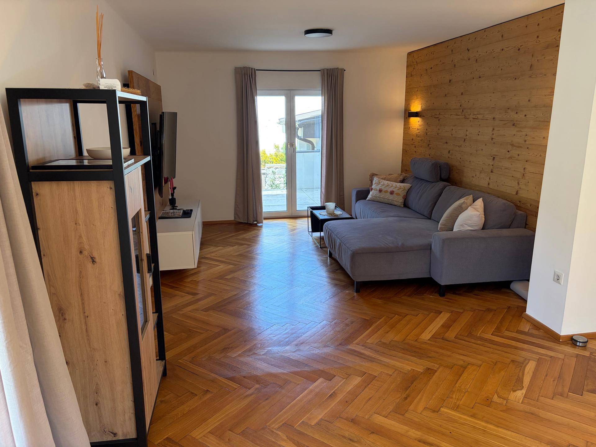 Gemütliches Wohnzimmer mit Fischgrätparkett, Holzwand und Zugang zur Terrasse.