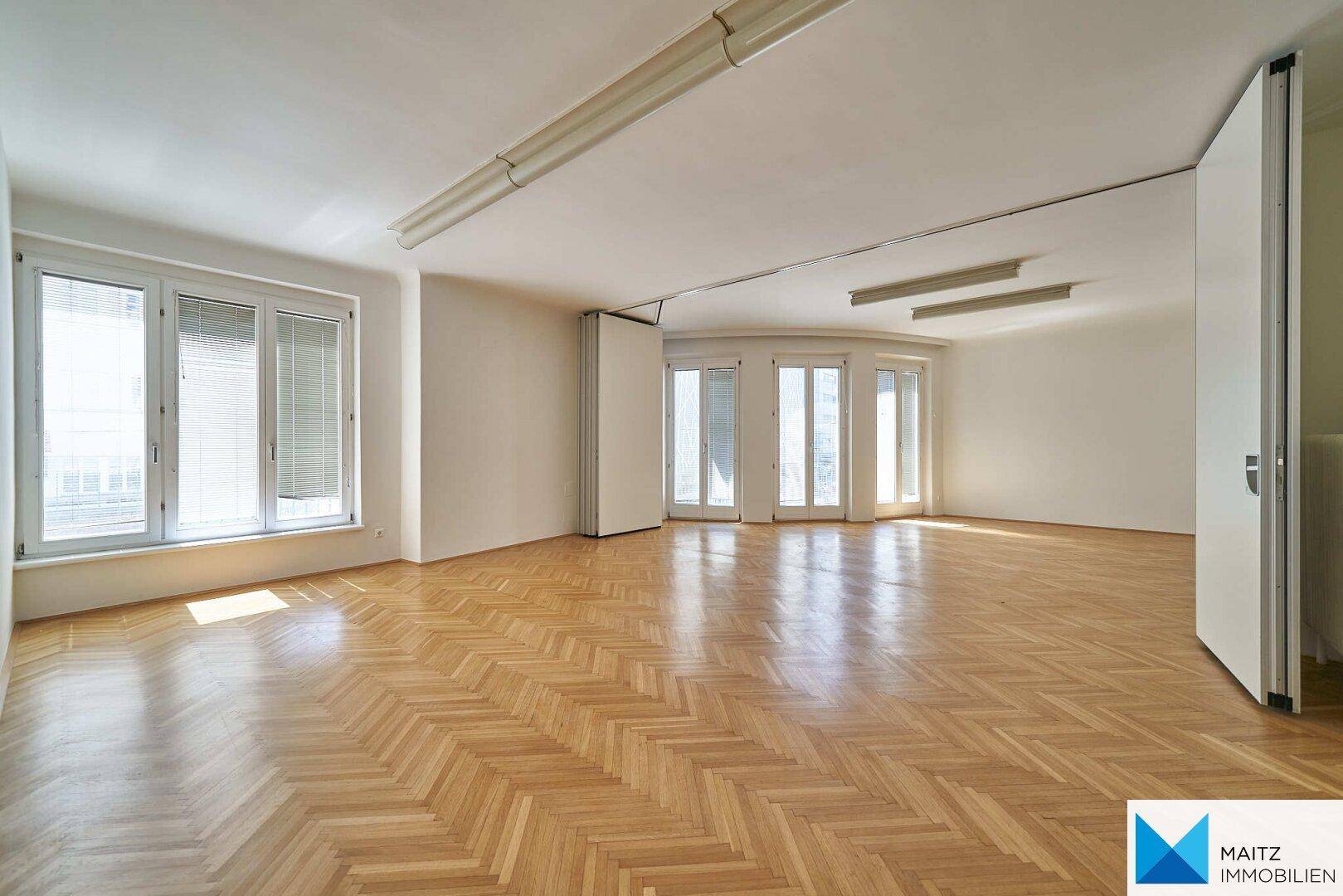 Heller Büroraum mit klassischem Fischgrätparkett und mehreren Fenstern für viel Tageslicht.