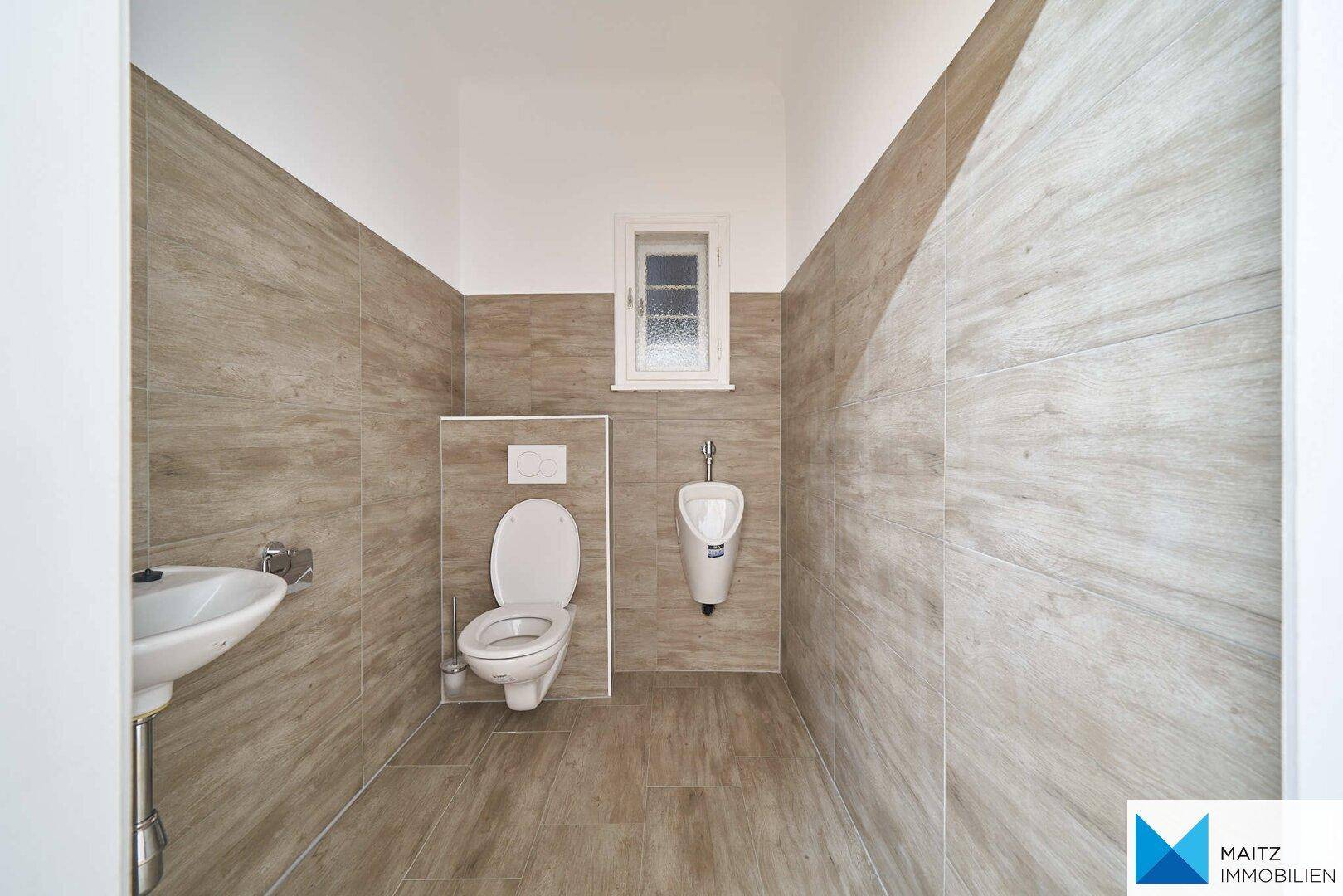 Modern gefliestes WC mit Toilette, Urinal, kleinem Waschbecken und einem Fenster.
