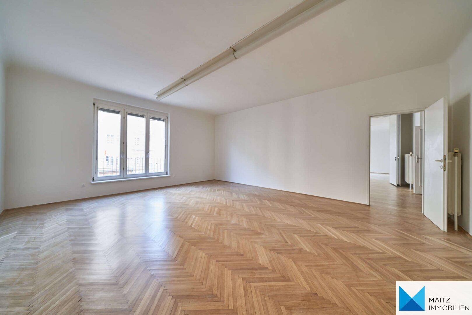 Großer, leerer Raum mit Fischgrätparkett, weiß gestrichenen Wänden und einem Fenster.