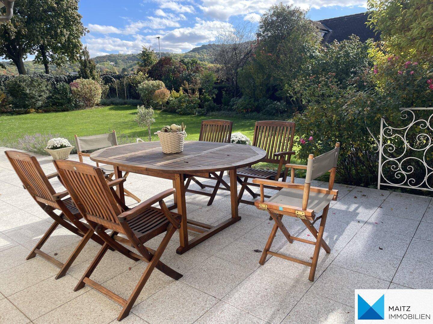 Sonnige Terrasse mit Holztisch und Stühlen, umgeben von üppigem Grün.